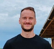 Smiling man with short brown hair and beard wearing a black shirt standing outdoors near a wooden structure.