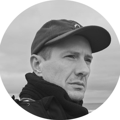 Black and white portrait of a man wearing a cap and scarf, looking to the side against a cloudy sky.