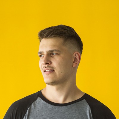 Young man with short dark hair, wearing a gray and black shirt, looking to his left against a yellow background.