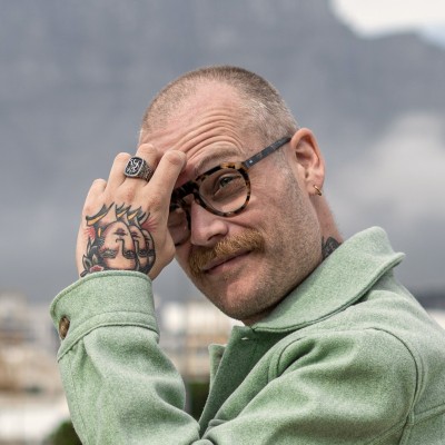 Man with glasses, mustache, earrings, and hand tattoo touching his forehead outdoors.