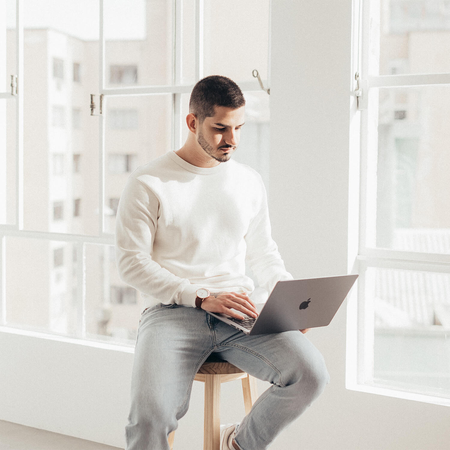 Man in white sweatshirt and jeans sitting on wooden stool working on a MacBook laptop by large windows.